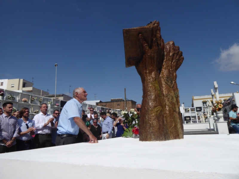 Un hombre en una ceremonia funeral, frente a una estatua de mano sosteniendo un libro, mientras un grupo de personas observa.