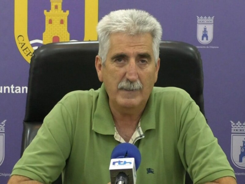 El hombre está sentado en una silla de oficina frente a un fondo azul con escudos heráldicos. Lleva una camiseta verde y tiene un micrófono en la mano izquierda, lo que sugiere una conferencia o entrevista.