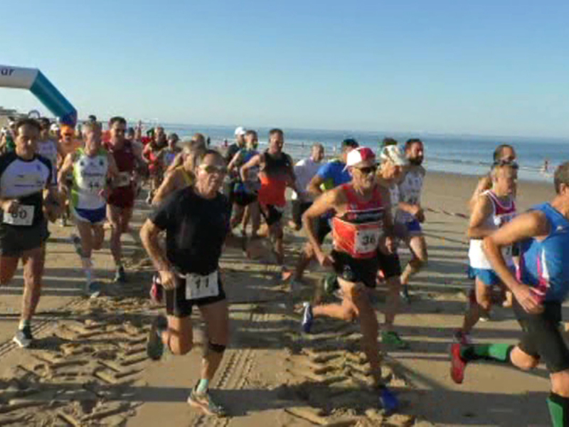 Corredores en una carrera de maratón en la playa, con el mar y el cielo azul al fondo.