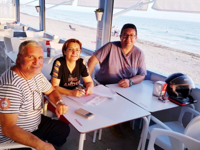 Tres personas sentadas en una mesa de playa, con un mar tranquilo y una arena clara al fondo. La persona en el centro lleva gafas de sol, mientras que la otra tiene un casco de motocicleta.