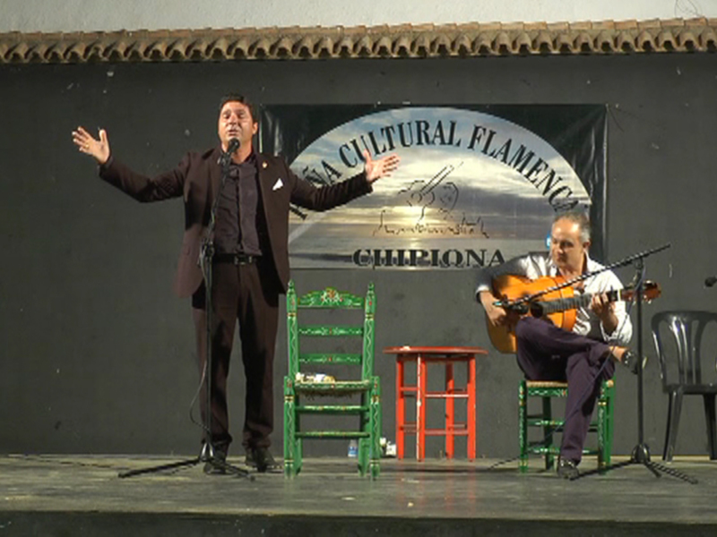 Un hombre en un traje negro se encuentra en el escenario, con un hombre tocando una guitarra en el fondo. La escena está iluminada y se sitúa en un escenario con un cartel que dice 'Cultura Flamenco'.
