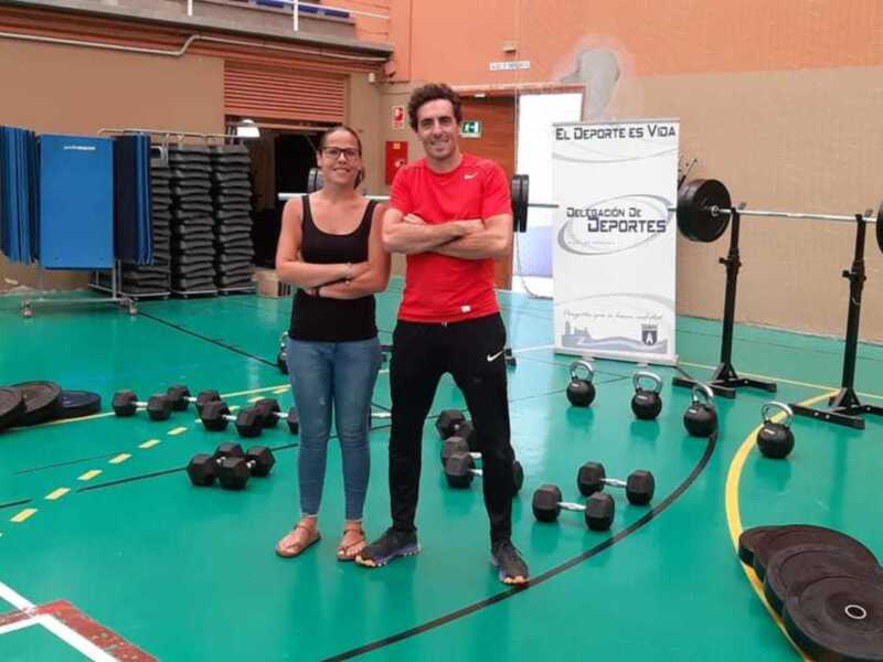En una sala de fitness, dos personas posan frente a un panel que dice "El deporte es vida". A la izquierda, una mujer con gafas y un top negro. A la derecha, un hombre en camiseta roja y pantalones negros. En el fondo, pesas y equipos de fitness.