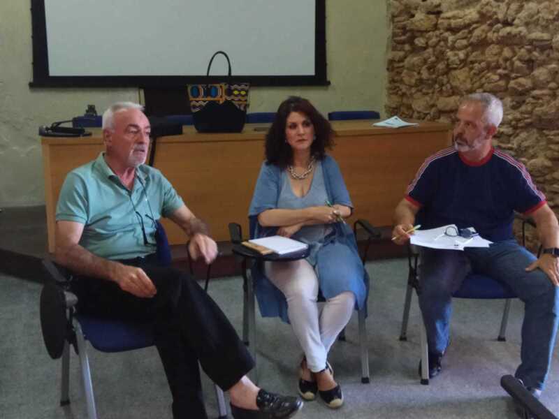 Tres personas sentadas en sillas, dos hombres y una mujer, frente a un fondo de paredes de ladrillo y una pantalla. La imagen parece ser de una reunión o conferencia en un entorno informal.