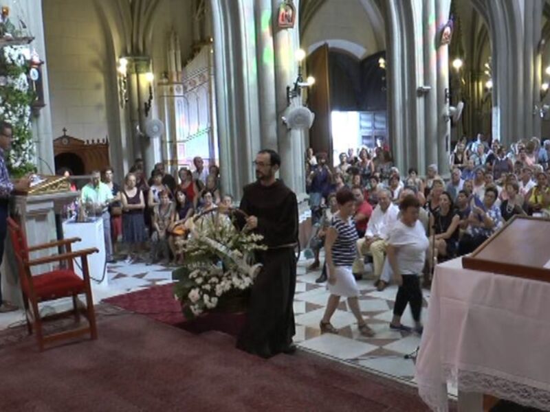 En una iglesia con arcos altos y columnas, un hombre en negro realiza gestos orales mientras una niña camina hacia él. La imagen está iluminada con luces de techo y una mesa con un papel en la esquina derecha.