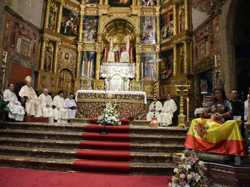 Interior de una iglesia con altar dorado, ofrendas florales y una bandera española en el frente.