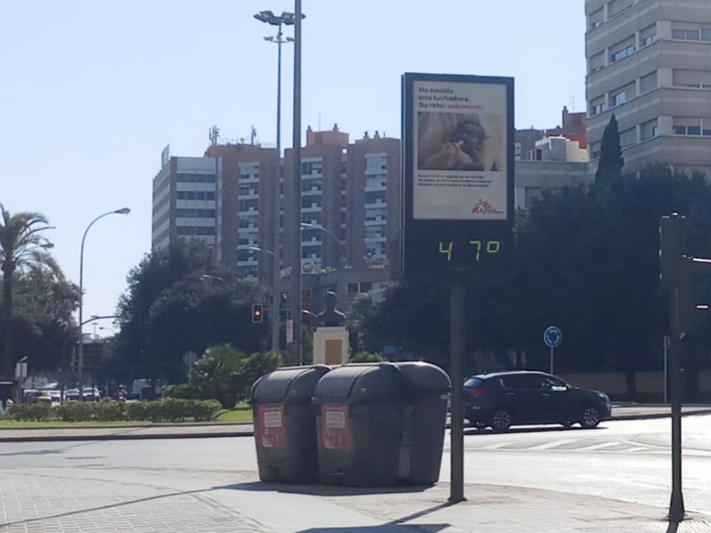 Se observa una calle urbana con edificios de varios pisos en el fondo. Hay dos contenedores de basura y un poste con una pantalla informativa que muestra información sobre el clima. La temperatura se indica como 47 grados Celsius. El cielo es claro y el horizonte está borroso debido a la distancia.