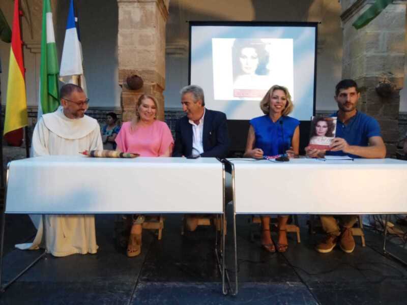 Se observa a una persona sentada en un escritorio, con un fondo de paredes de ladrillo y una pantalla proyectando una imagen. La persona está sosteniendo un objeto en sus manos, posiblemente un libro o una revista. La escena parece ser parte de una presentación o conferencia, dada la presencia del escritorio y la pantalla.