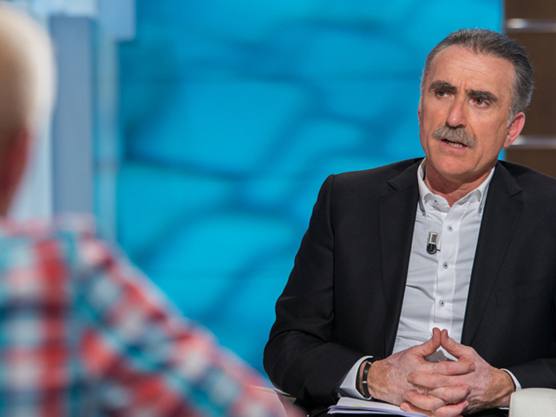 Dos hombres conversan en un estudio de televisión, uno con una camisa a cuadros y el otro con una chaqueta negra. El fondo es azul con líneas horizontales.
