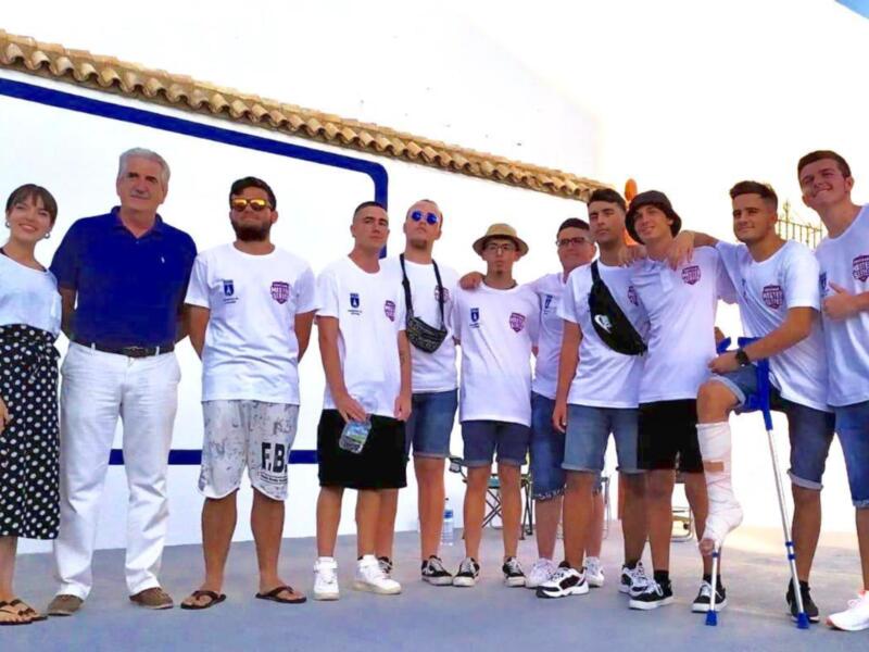 Una imagen de un grupo de personas posando frente a una pared blanca con detalles azules. La mayoría están vestidos con camisetas blancas y pantalones cortos, algunos con faldas. La persona de la izquierda lleva un vestido negro a cuadros y sandalias. En el fondo, se ve una estructura de ladrillo con un techo de tejas.