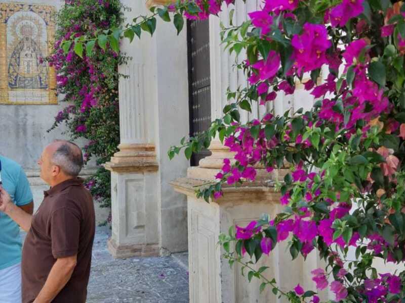 Un hombre con una camiseta azul está entrevistando a otro hombre en un entorno de arquitectura tradicional con flores coloridas al fondo.