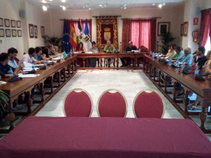 Una sala de conferencias con una mesa central cubierta por un mantel rojo, rodeada por sillas y mesas. Hay personas sentadas en las sillas, algunas de ellas escribiendo o hablando. La pared está decorada con banderas y cuadros.