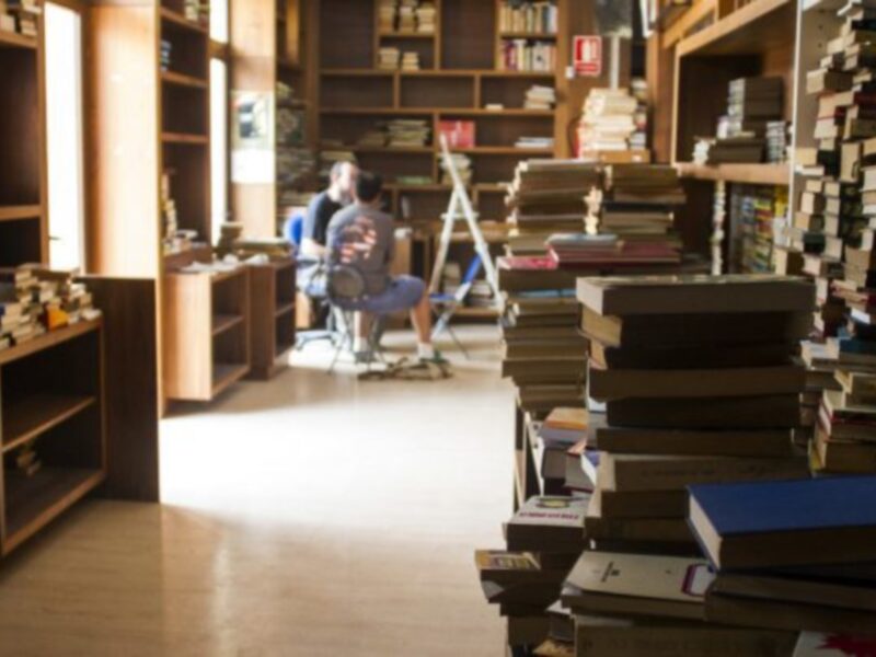 Un hombre está sentado en un sillón de pie, rodeado por una gran cantidad de libros apilados. La habitación está llena de estanterías llenas de libros, creando un ambiente acogedor y lleno de conocimiento.