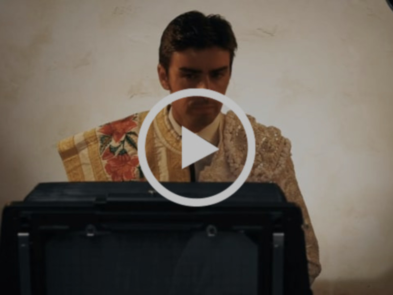Un hombre en una habitación con una camisa de lana beige y un chaleco floral, mirando hacia la cámara.