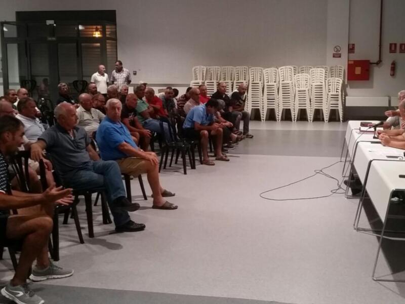 Una sala de conferencias con asistentes sentados en sillas alrededor de una mesa. La pared está llena de pantallas y el suelo es claro.