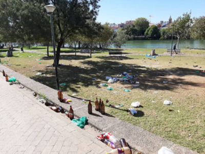 Área de parque con basura dispersa en el suelo y en el borde de la acera, cerca de un lago y árboles.