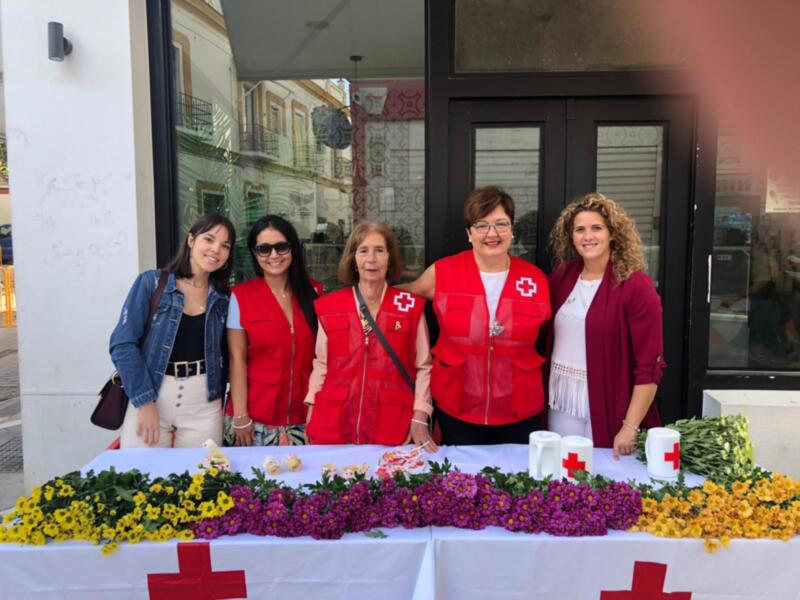 Las voluntarias de Cruz Roja posan frente a una mesa decorada con flores y banderas rojas, en un entorno urbano.