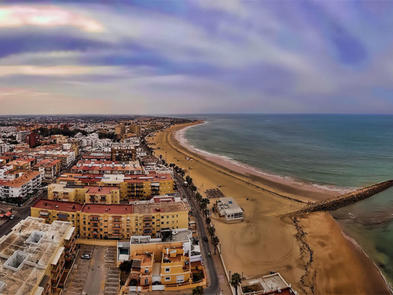Vista panorámica de una ciudad costera con edificios de apartamentos y una playa arenosa bordeada por un dique de piedra.
