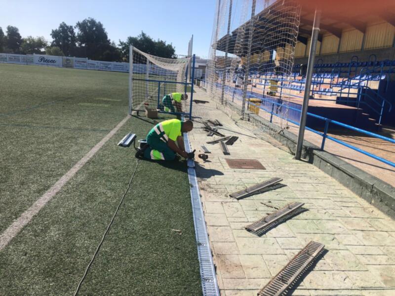 Un trabajador en un uniforme verde está ajustando una valla de metal en un campo de fútbol artificial. La imagen muestra detalles como el césped verde, la valla de metal y una parte del estadio con asientos azules.