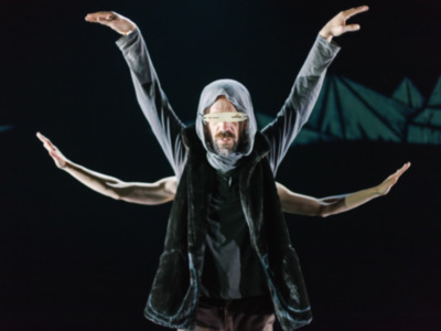 Dancer in a hooded outfit with arms outstretched, performing on stage with a dramatic backdrop.