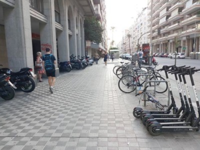 En una calle de la ciudad, varias motocicletas y bicicletas están aparcadas al lado de un edificio con ventanas grandes. Al fondo, se pueden ver algunos coches estacionados y una parada de autobús. La imagen muestra un ambiente urbano con edificios altos y una calle adoquinada.