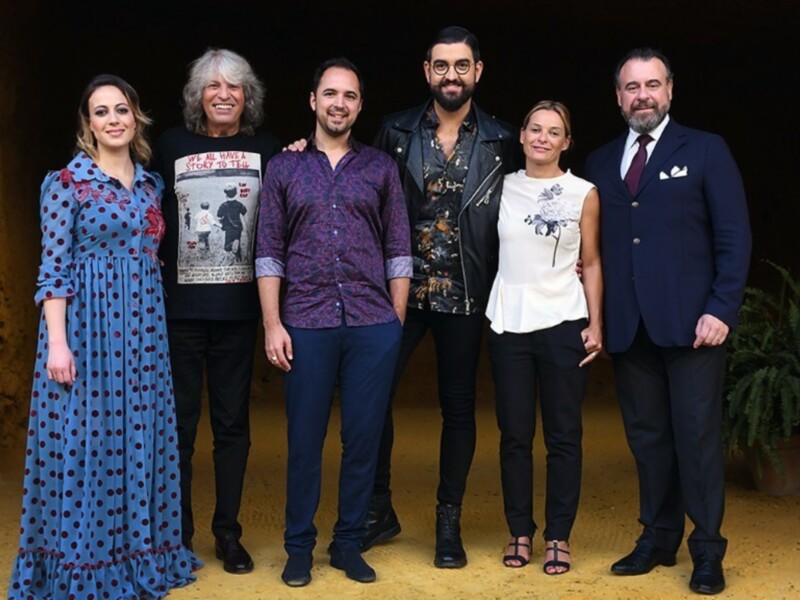 Una imagen de cinco personas posando frente a un fondo de paredes amarillas. La persona en el centro lleva una camiseta con letras negras y la derecha tiene un traje oscuro. La izquierda muestra una camiseta con letras blancas y la derecha, un traje azul oscuro.