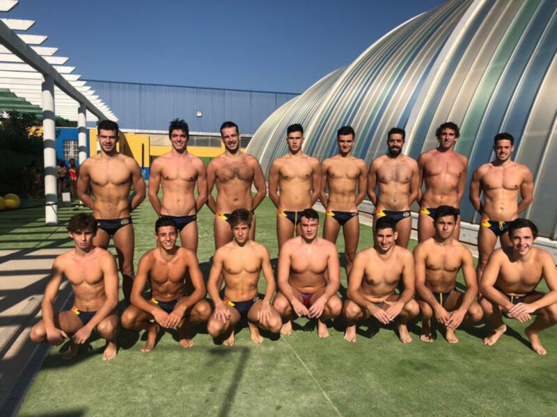 Un grupo de jóvenes hombres posa en una cancha de fútbol, todos desnudos excepto por sus bañadores. El fondo muestra una estructura metálica curva y un cielo azul claro, sugiriendo un entorno al aire libre.