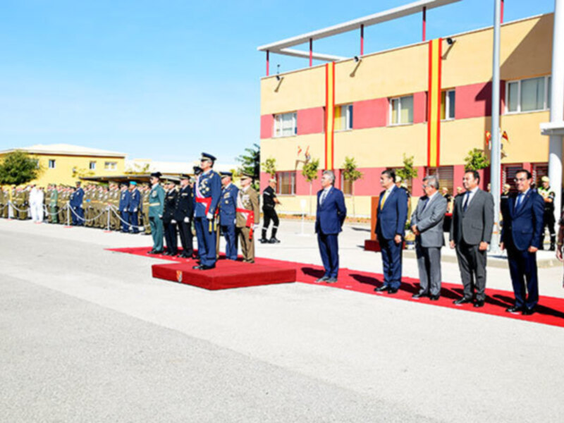 Una ceremonia militar en un edificio amarillo con detalles rojos y azules, donde militares y civiles se encuentran en uniforme. La imagen muestra una fila de personas, algunas en trajes militares y otras civiles, posando para la foto. La escena está iluminada por el sol y parece ser en un entorno urbano con edificios al fondo.