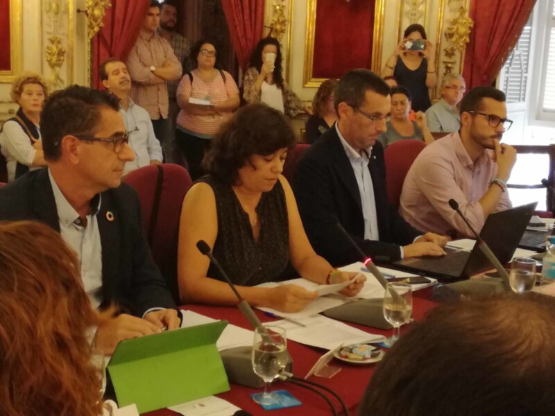 Una reunión formal en un salón de conferencias con personas sentadas alrededor de una mesa, sosteniendo documentos y usando laptops. La decoración incluye cortinas rojas y un marco dorado en el fondo, creando un ambiente de trabajo serio.