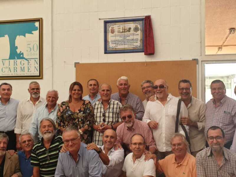 Grupo de personas posando para una foto en un salón de reuniones. En el fondo, se puede ver una pared con un cartel que dice 'VIRGEN del CARMEN' y una placa con texto en español.
