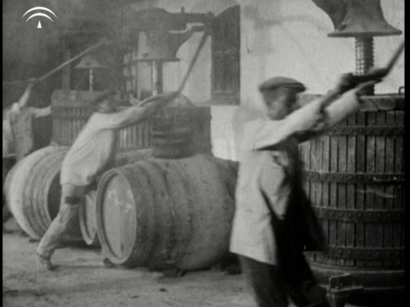 Imagen antigua de trabajadores arrastrando barriles grandes en un entorno industrial.