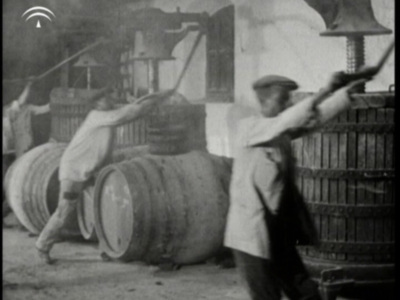 Imagen antigua de trabajadores arrastrando barriles grandes en un entorno industrial.