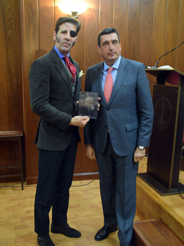 Dos hombres en trajes formales, uno sosteniendo una foto antigua, posan frente a un fondo de madera y un micrófono en un ambiente formal.