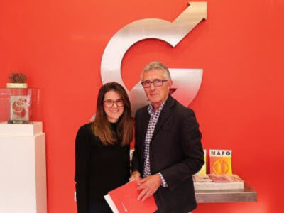 Dos personas posan frente a una pared roja con un gran logotipo de Google. La persona de la izquierda lleva una camisa negra y gafas, mientras que la de la derecha usa un traje negro con una camisa a cuadros. En el fondo, se pueden ver algunos objetos sobre un mostrador blanco.