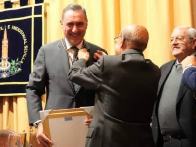 Se observa a un grupo de personas celebrando, posiblemente en una ceremonia o evento formal. Al fondo hay un escudo con texto y una bandera, lo que sugiere que podría ser un lugar de importancia o institución. La celebración parece ser en un ambiente formal, posiblemente dentro de una sala o auditorio.