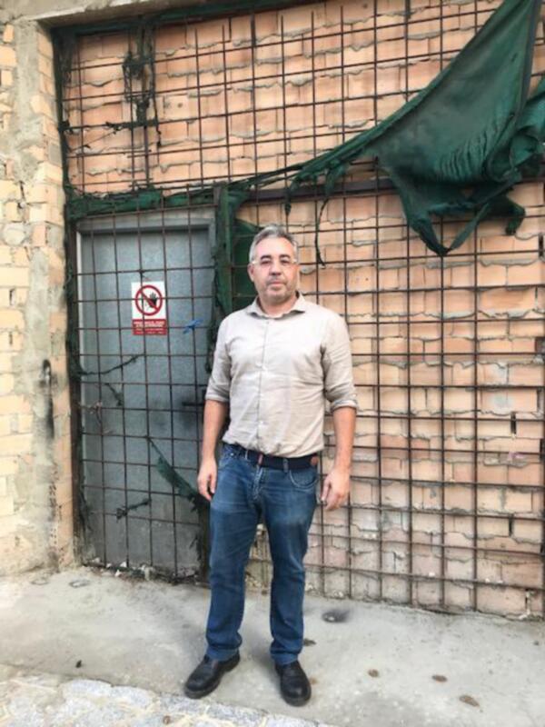 Un hombre de mediana edad, con barba y cabello gris, posa frente a una puerta metálica de un edificio con paredes de ladrillo. Lleva una camisa beige, jeans y zapatos negros. La puerta tiene un letrero rojo con una cruz dentro de un círculo, indicando prohibido entrar. La imagen parece ser de una zona urbana.