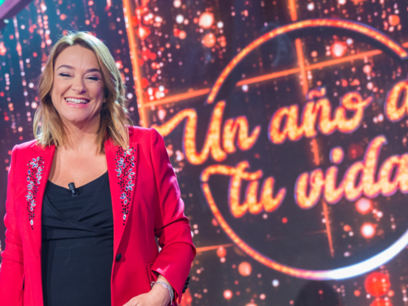 A una presentadora sonriente en un escenario iluminado con luces rojas y azules, frente a una pantalla que dice "Un año de tu vida". La presentadora lleva un traje rojo con detalles en la manga y una camisa negra.