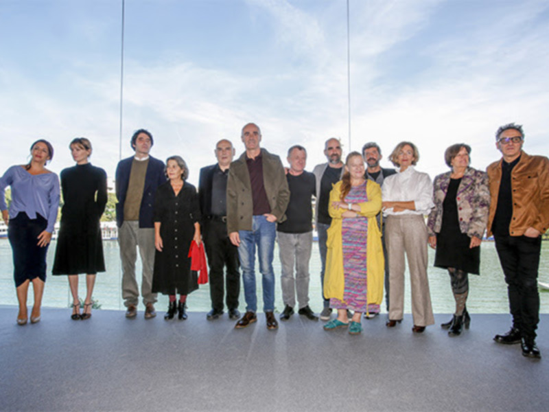 Una fila de diez personas posando frente a un marco de cristal que refleja el cielo azul y la bahía. La imagen captura una reunión formal o profesional en un entorno urbano con vistas al agua.