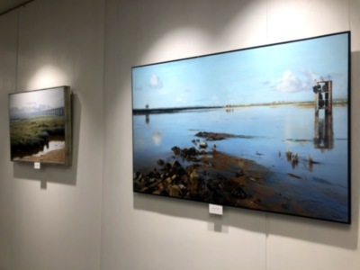 Dos cuadros de arte en una pared, uno grande y otro pequeño. El gran cuadro muestra un paisaje costero con agua azul, cielo azul y una estructura de madera en el horizonte. El cuadro pequeño muestra un paisaje similar pero más oscuro y con menos detalles visibles.