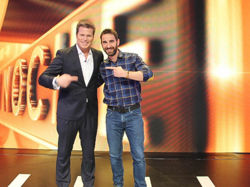 Dos hombres en una escena de televisión, uno con traje y el otro con camisa a cuadros, posando con los brazos extendidos. El fondo es una pantalla con luces y una iluminación naranja.
