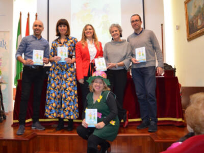 Una familia feliz sostiene libros infantiles en un evento educativo. La imagen muestra a padres y sus hijos, todos sonrientes y orgullosos de sus logros. La escena se desarrolla en un salón con una pantalla proyectando un diseño floral, y una bandera visible. La iluminación es cálida y el ambiente parece ser un ambiente de celebración o reconocimiento.