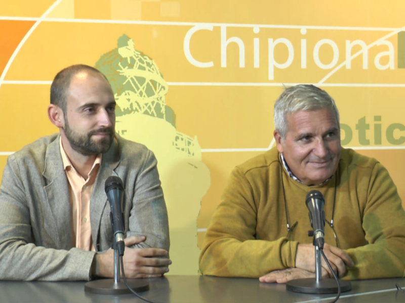 Dos hombres en una conferencia de prensa, con un fondo amarillo y letras que dicen "Chipioniana Hoy".
