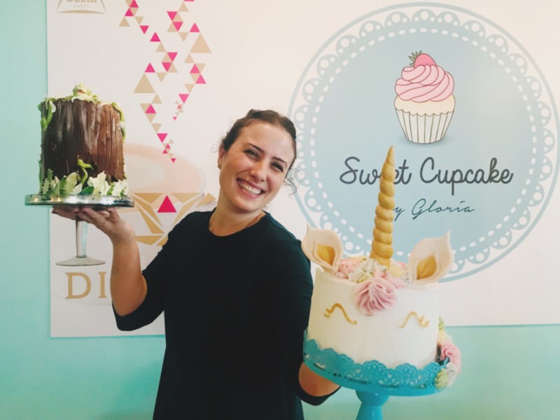 A una persona sosteniendo un pastel de unicornio con detalles en color rosa y dorado. El fondo tiene una decoración geométrica de triángulos en colores pastel y un logotipo que dice "Sweet Cupcake".