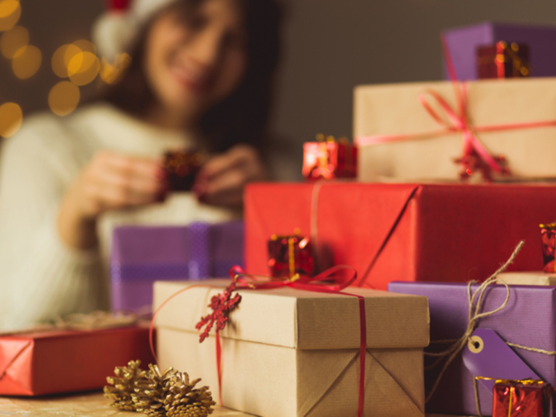 Una mujer sonriendo, con una gorrita de Navidad en la cabeza, frente a un montón de regalos envueltos con cintas rojas y doradas, en un ambiente festivo con luces de Navidad difuminadas al fondo.