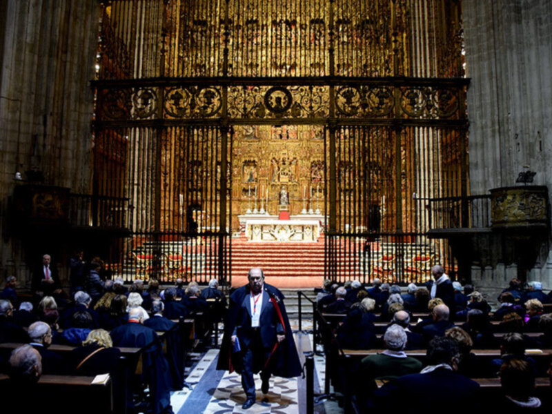 Interior de una iglesia con una gran arquitectura en el fondo, donde una persona con una capa negra y un cetro rojo se dirige a una multitud sentada en los bancos. El altar es el centro de atención con una gran imagen en el fondo.
