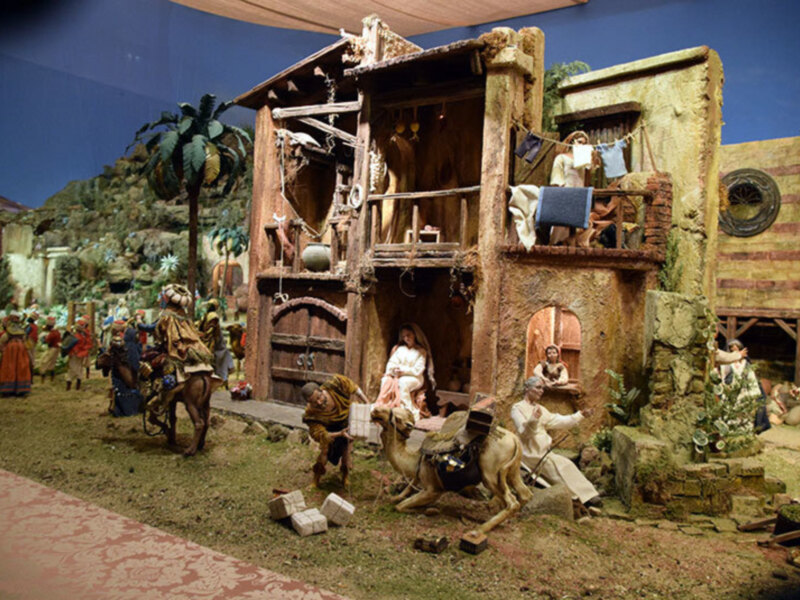 Un escenario de natividad con figuras humanas y animales en una representación de la natividad de Navidad, con detalles como una casa de madera, un camello y figuras vestidas de blanco y rojo.