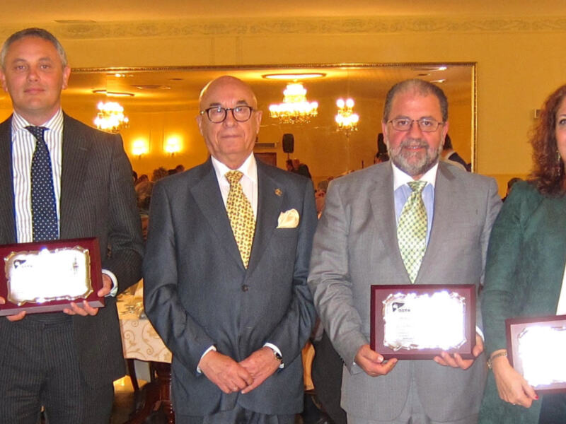 Grupo de personas en un evento formal, todos vestidos con trajes y corbatas. Algunos sostienen reconocimientos o medallas, lo que sugiere un ambiente de celebración o premiación. La decoración y la iluminación indican que se trata de un evento en una sala o salón.