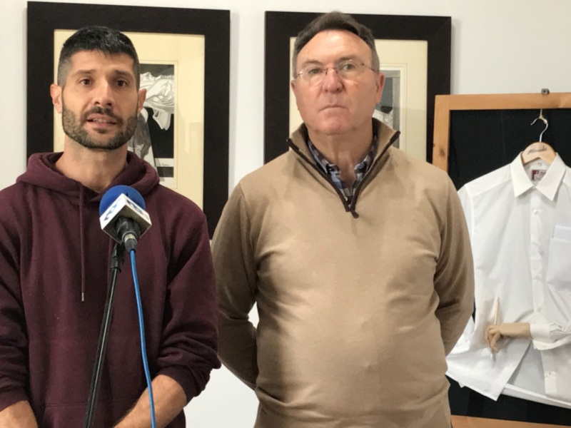 Dos personas en una reunión, uno con un micrófono y el otro con una camisa blanca en la mano.