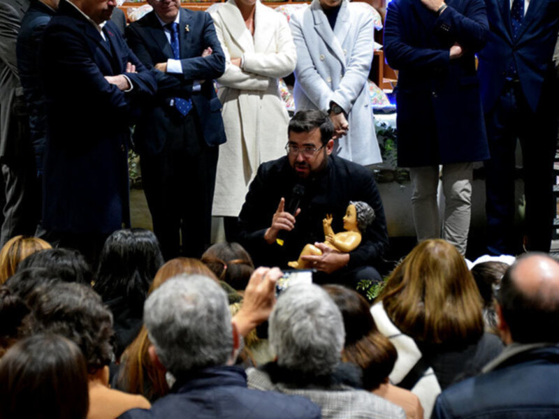 Una figura de una niña con un vestido de noche y un sombrero se encuentra en el centro de una reunión formal, rodeada por personas vestidas con trajes y abrigos. La escena sugiere una ceremonia o evento importante.