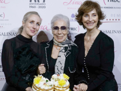Tres mujeres sonrientes posan con pasteles en un evento celebrativo. El fondo es una pantalla con logotipos y texto que parece ser parte de un aniversario.