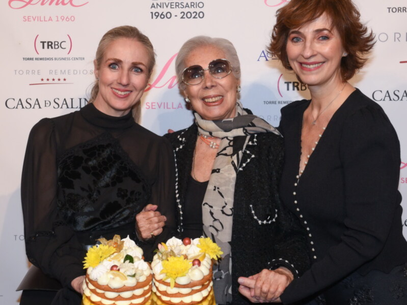 Tres mujeres sonrientes posan con un pastel de cumpleaños en un evento de celebración de 60 años en Sevilla, con logotipos de empresas y eventos en el fondo.